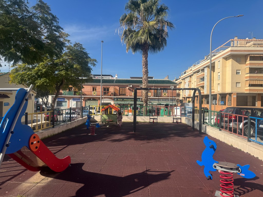 A playground in Málaga featuring colorful play equipment, including a slide and spring riders, surrounded by palm trees and residential buildings under a clear blue sky.